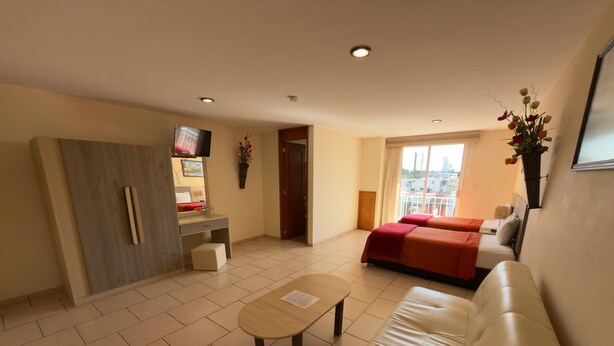 Ein modernes Hotelzimmer mit einer Couch, einem kleinen Tisch, einem Schreibtisch mit Stuhl, einem Bett mit roter Bettwäsche, einem Fernseher und einem Fenster mit Blick auf die Stadt.