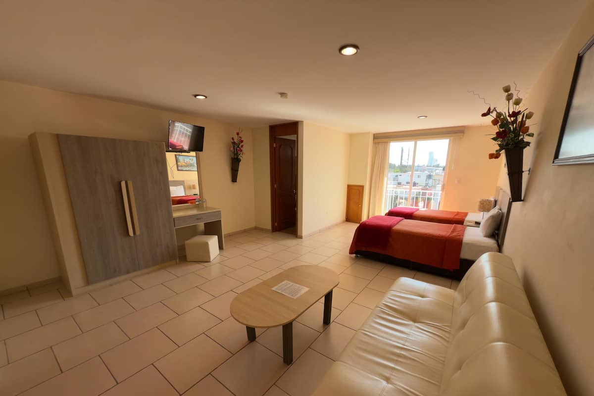 A modern hotel room with a sofa, a small table, a desk with a chair, a bed with red bedding, a TV, and a window with a view of the city.