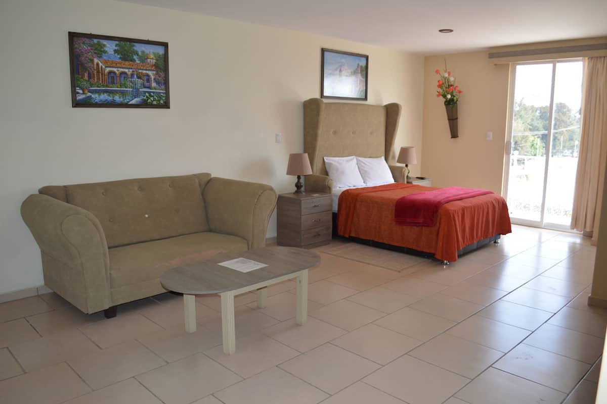A hotel room with a sofa, a small table, a bed with a red bedspread, a nightstand with lamps, and a painting on the wall.