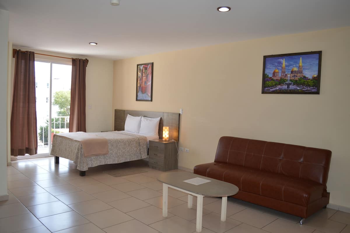 A hotel room with a bed, a sofa, a nightstand with a lamp, and two paintings on the wall.