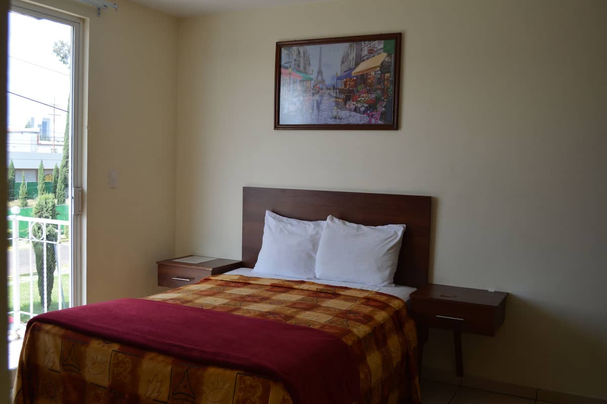 A hotel room with a bed, a nightstand, a bedside table, a window with a view of buildings, and a framed picture on the wall.