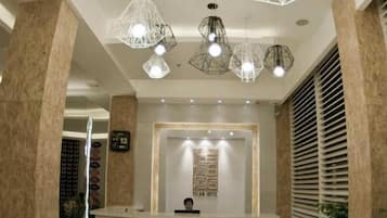 Zona de recepció d'hotel amb un disseny modern, que inclou un taulell de recepció, il·luminació penjant geomètrica i un gran finestral amb persianes.