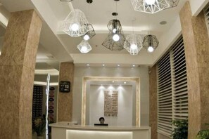A hotel reception area with a modern design, featuring a reception desk, geometric pendant lighting, and a large window with blinds. - Yilan Hotel (Wuyishan)