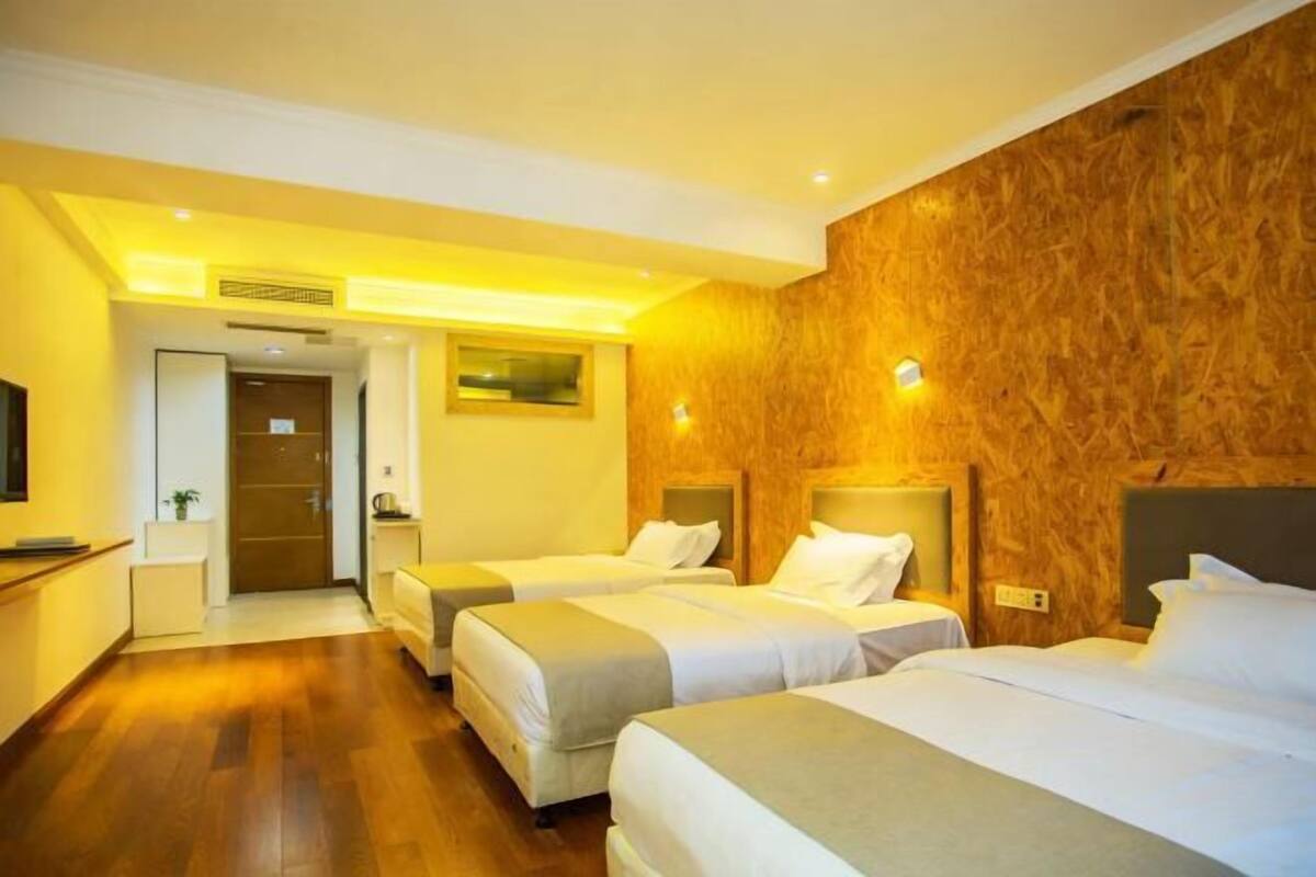 A hotel room with three beds, wooden paneling on one wall, a small desk with a chair, and a door leading to another room.