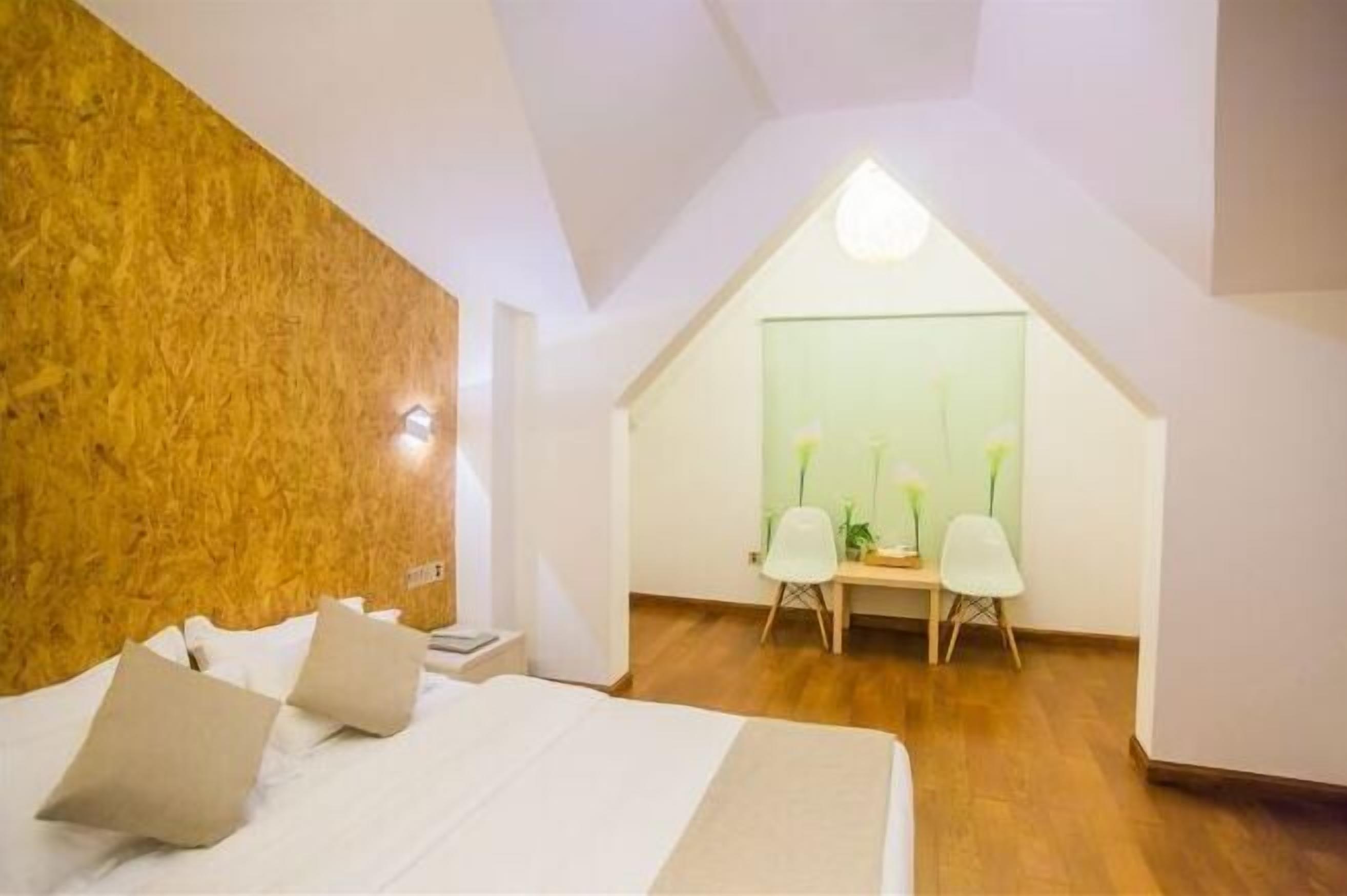 A bedroom with a sloped ceiling, a bed with pillows, a desk with chairs, and a green wall with a window.