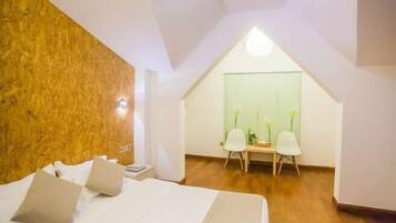 A bedroom with a sloped ceiling, a bed with pillows, a desk with chairs, and a green wall with a window.
