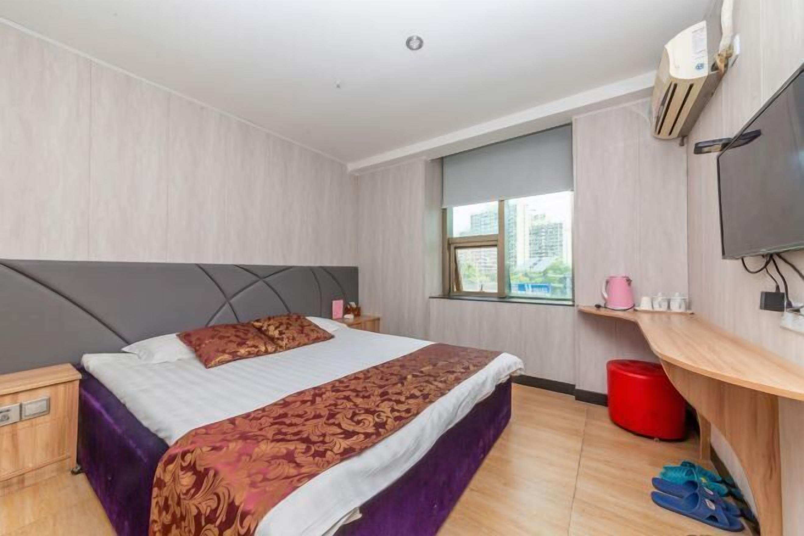 Una camera d'albergo con un letto, una TV, una finestra, un cestino rosso e una mensola in legno con vari oggetti.