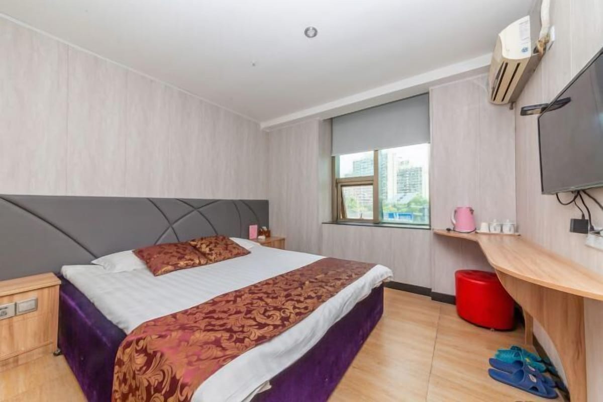 Une chambre d’hôtel avec un lit, une télévision, une fenêtre, une poubelle rouge et une étagère en bois sur laquelle sont posés divers objets.