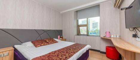 Habitación de hotel con cama, televisor, ventana, un cubo de basura rojo y estante de madera con objetos.