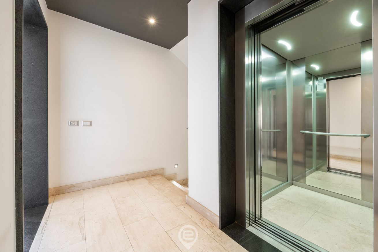 Interior moderno com porta de elevador em vidro, piso ladrilhado e parede lisa com interruptor de luz.