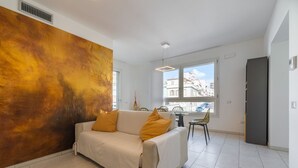 A modern living room with a white sofa, a large abstract painting, a dining area with chairs, and a view of buildings through the window. - Ada Bright Escape Apartment (Cagliari)