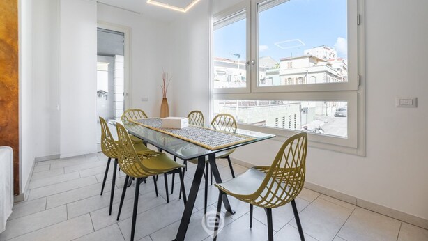 A modern dining area with a glass table and yellow chairs, large windows with a city view, and a geometric light fixture. - Ada Bright Escape Apartment (Cagliari)
