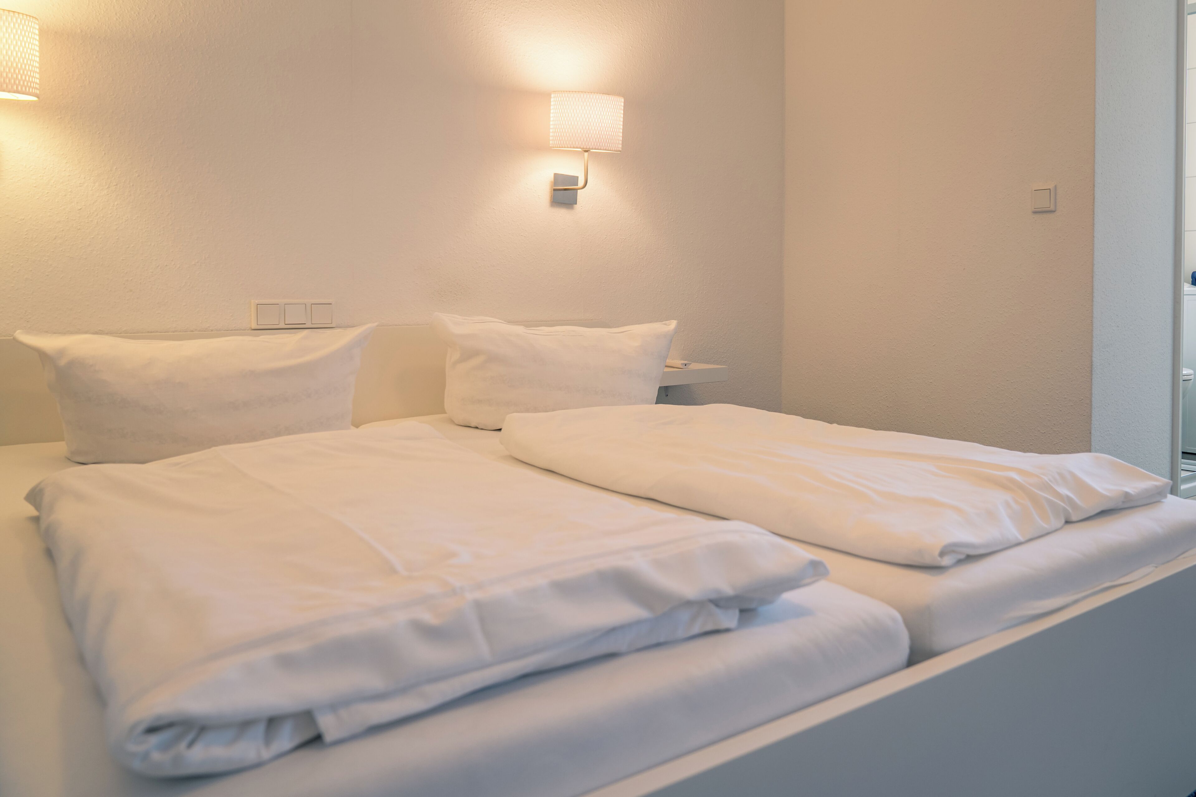 A double bed with white linens, a bedside table, and a wall-mounted lamp.