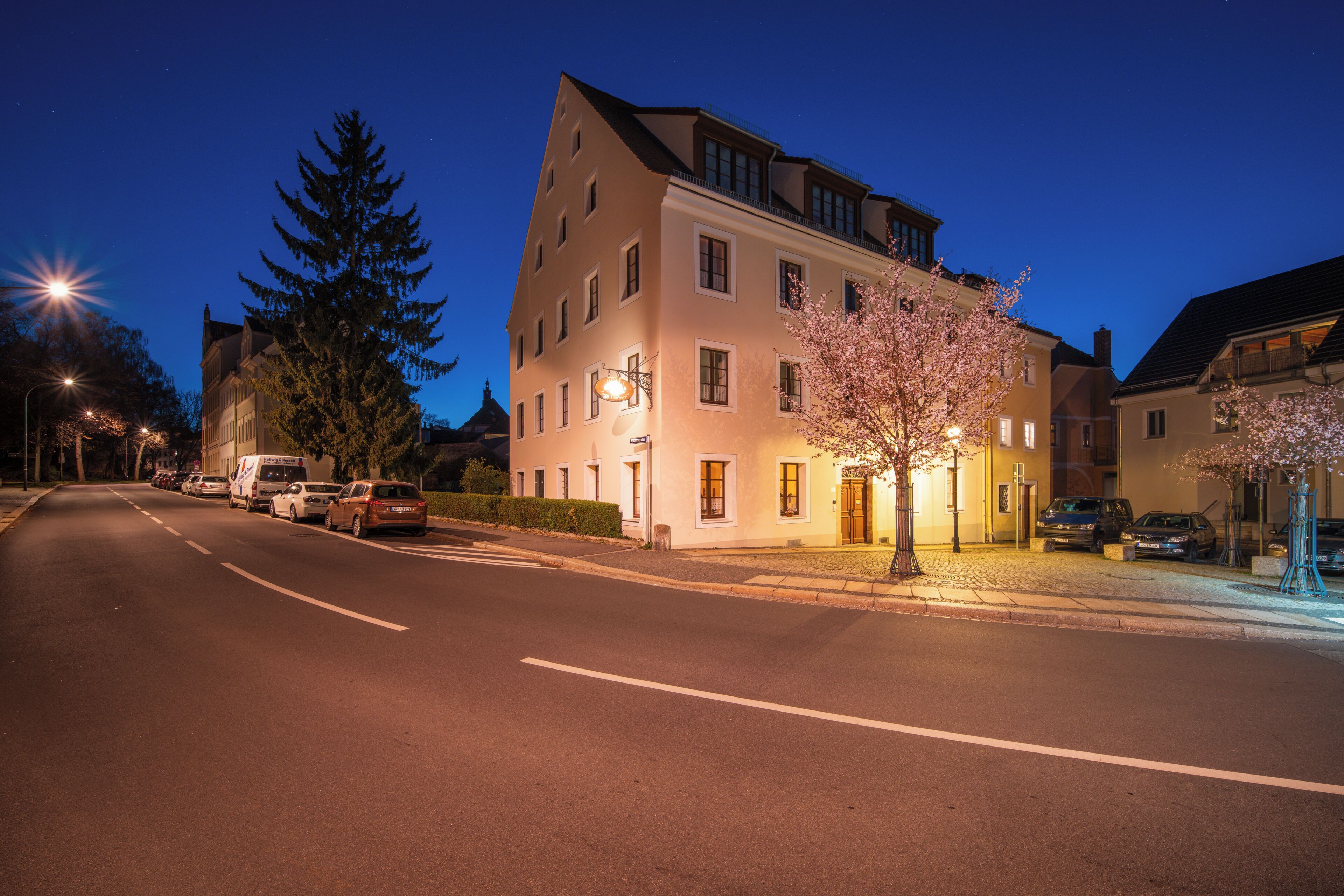 A multi-story building with a well-lit entrance, parked cars, and a tree with pink blossoms.