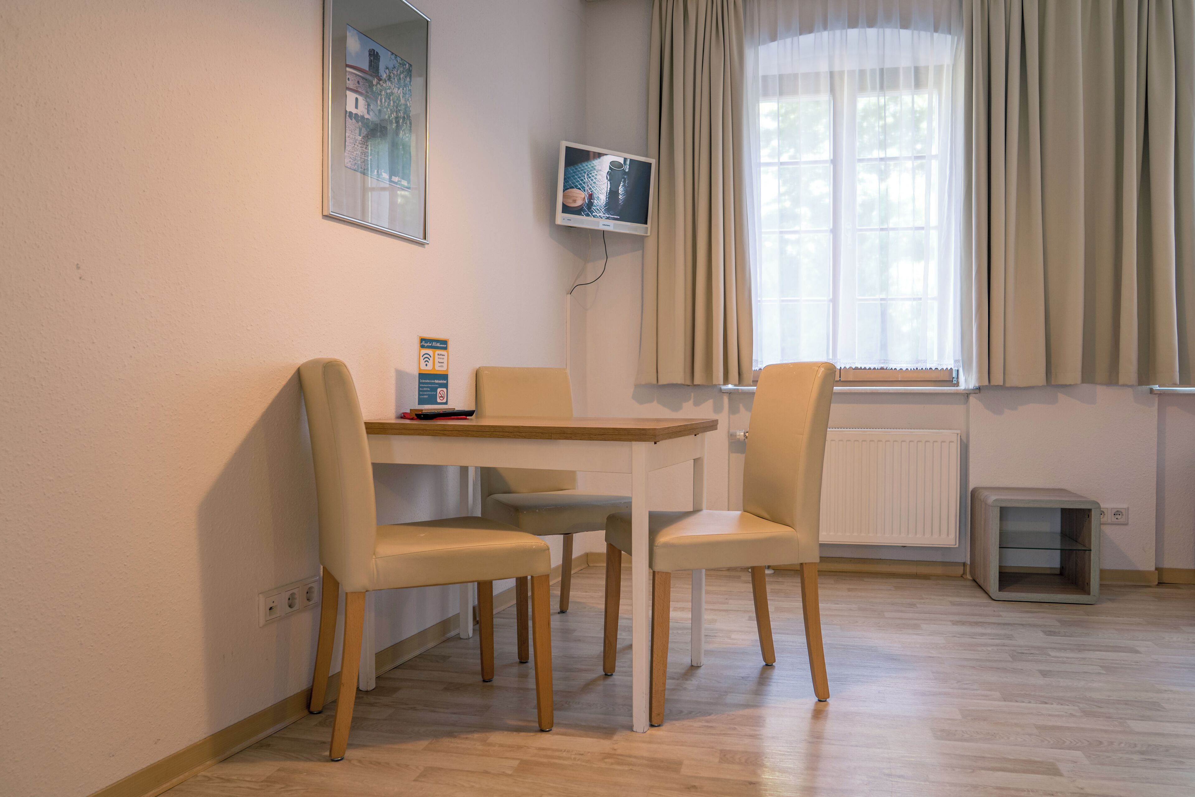 A dining area with a table and four chairs, a television on the wall, a window with curtains, and a radiator.