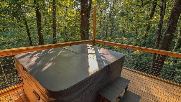A cozy outdoor seating area with a cushioned bench and a table, surrounded by a wooden deck and a wire fence, overlooking a dense forest.