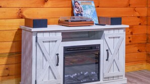 A wooden cabin interior with a mounted television displaying a landscape, a small table with a picture frame, and a fireplace.