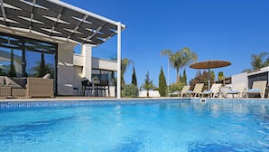 A modern pool area with lounge chairs, a dining table, and a covered seating area. - Villa Camellia - 3bed Bungalow large pool (Protaras)