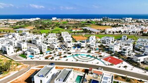A modern residential area with white buildings, a central circular plaza, and a view of the sea. - Villa Camellia - 3bed Bungalow large pool (Protaras)