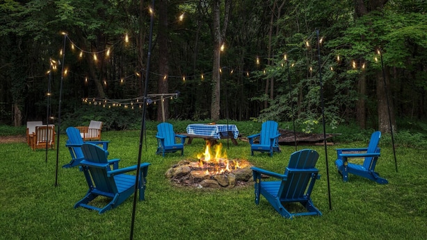 A fire pit surrounded by blue Adirondack chairs in a forested area with string lights. - 4BR 50-Acre Ridge Retreat | Nature & Fire Pit (Greensboro)