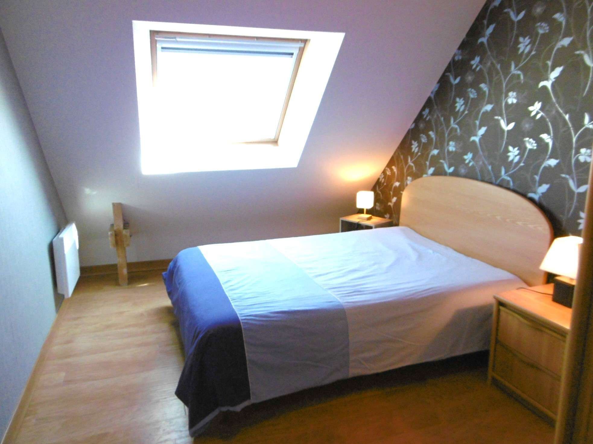 Una habitación pequeña con cama individual, techo inclinado, tragaluz, mesita de noche con lámpara y pared con estampado floral.