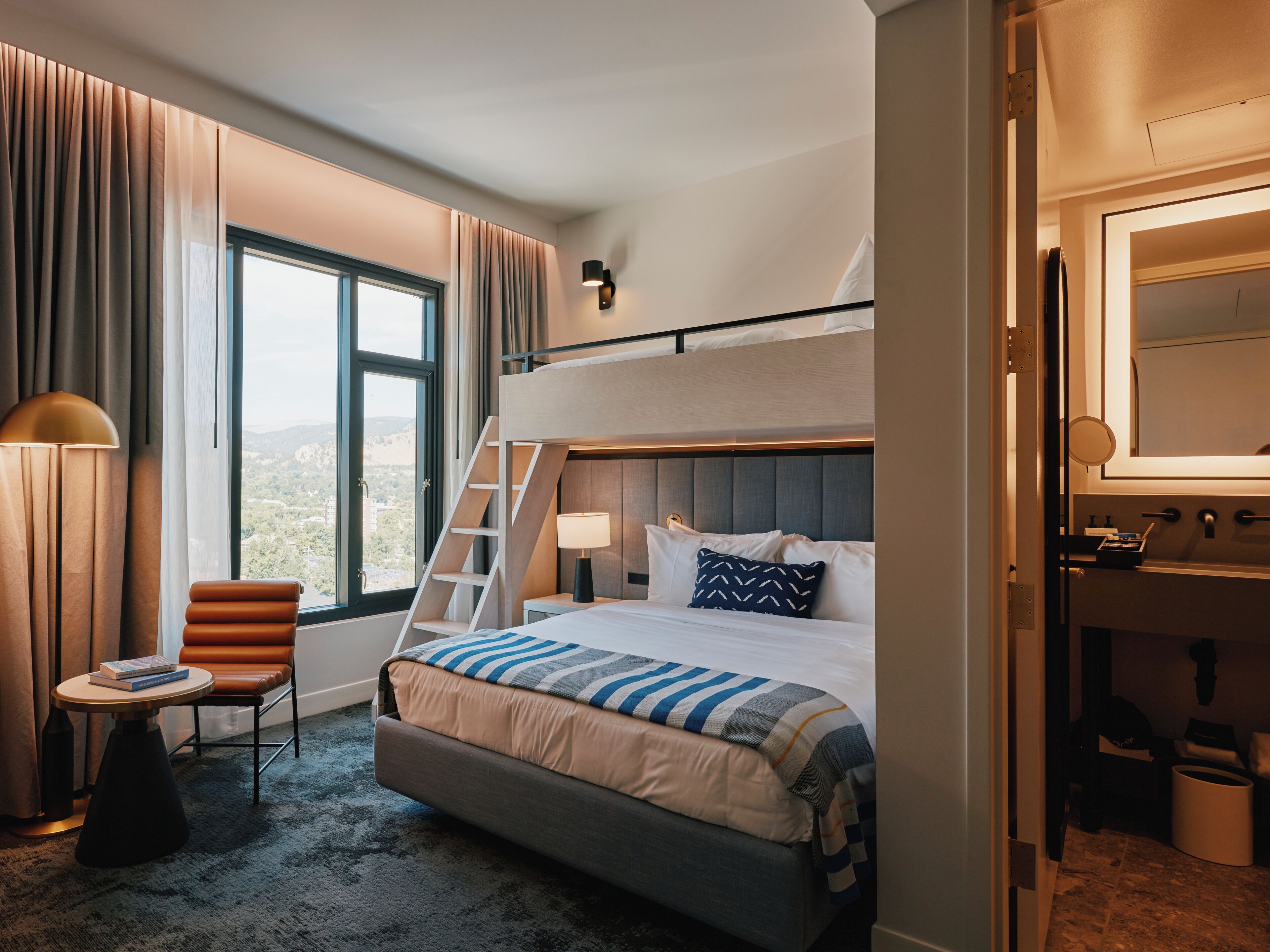 A hotel room with a bunk bed, a desk with a chair, a window with curtains, and a bathroom visible through an open door.