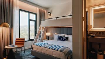 A hotel room with a bunk bed, a desk with a chair, a window with curtains, and a bathroom visible through an open door.