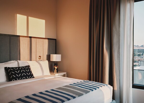 A hotel room with a large bed, a nightstand with a lamp, and a view of a cityscape through the window. - Limelight Boulder (Boulder)
