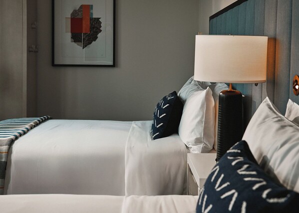A hotel room with two beds, a bedside table with a lamp, and a framed artwork on the wall.