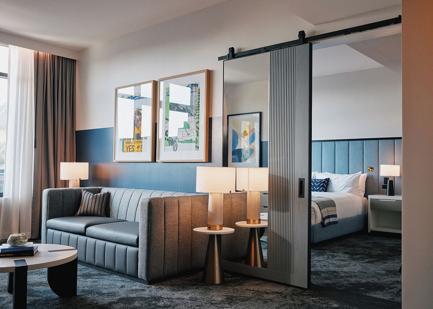 A modern hotel room with a sofa, two bedside tables with lamps, a bed with a headboard, and two framed artworks on the wall.