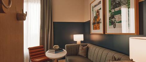 Habitación de hotel con sofá, una pequeña mesa redonda con libros, una silla, una lámpara y dos cuadros en la pared.