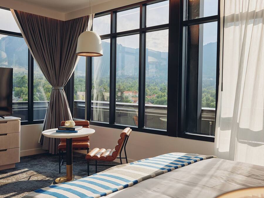 A hotel room with a large window, a bed, a desk, a chair, and a lamp.