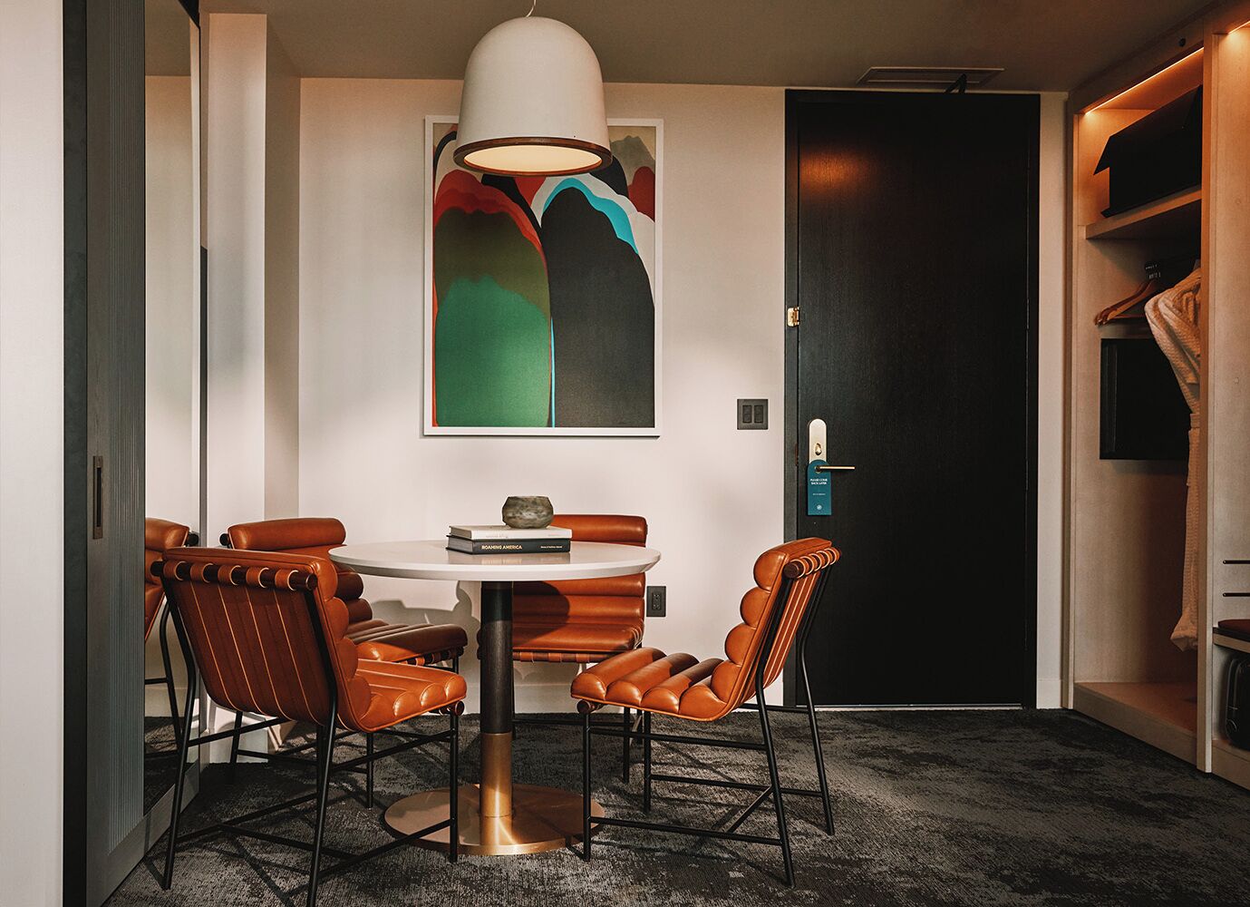 Un área de comedor con una mesa redonda, sillas de cuero, una lámpara colgante y un cuadro abstracto de colores en la pared.