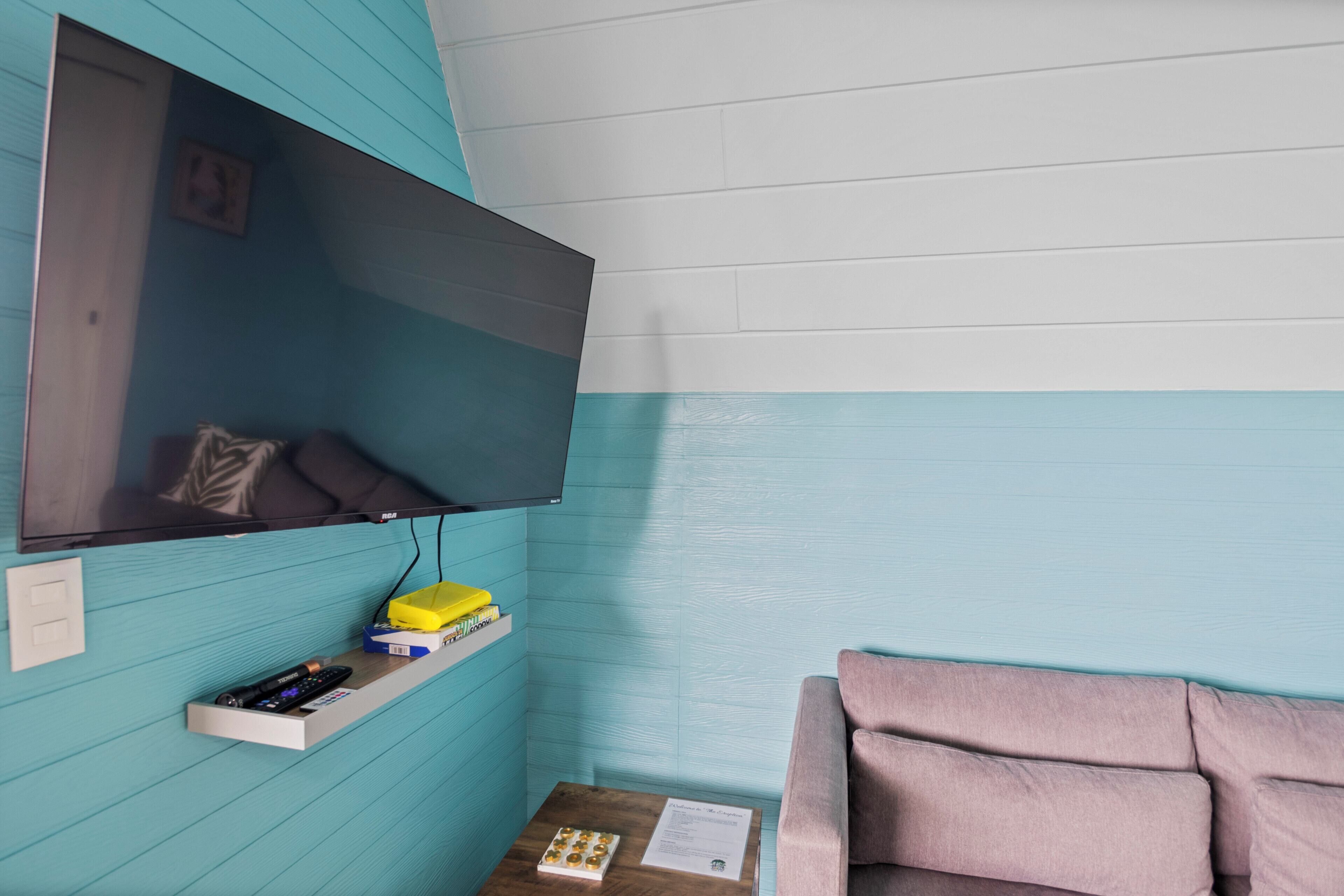 A room with teal walls, a mounted TV, a wooden side table with a remote control, a pink couch, and a shelf with books and a yellow box.