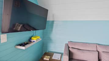A room with teal walls, a mounted TV, a wooden side table with a remote control, a pink couch, and a shelf with books and a yellow box.