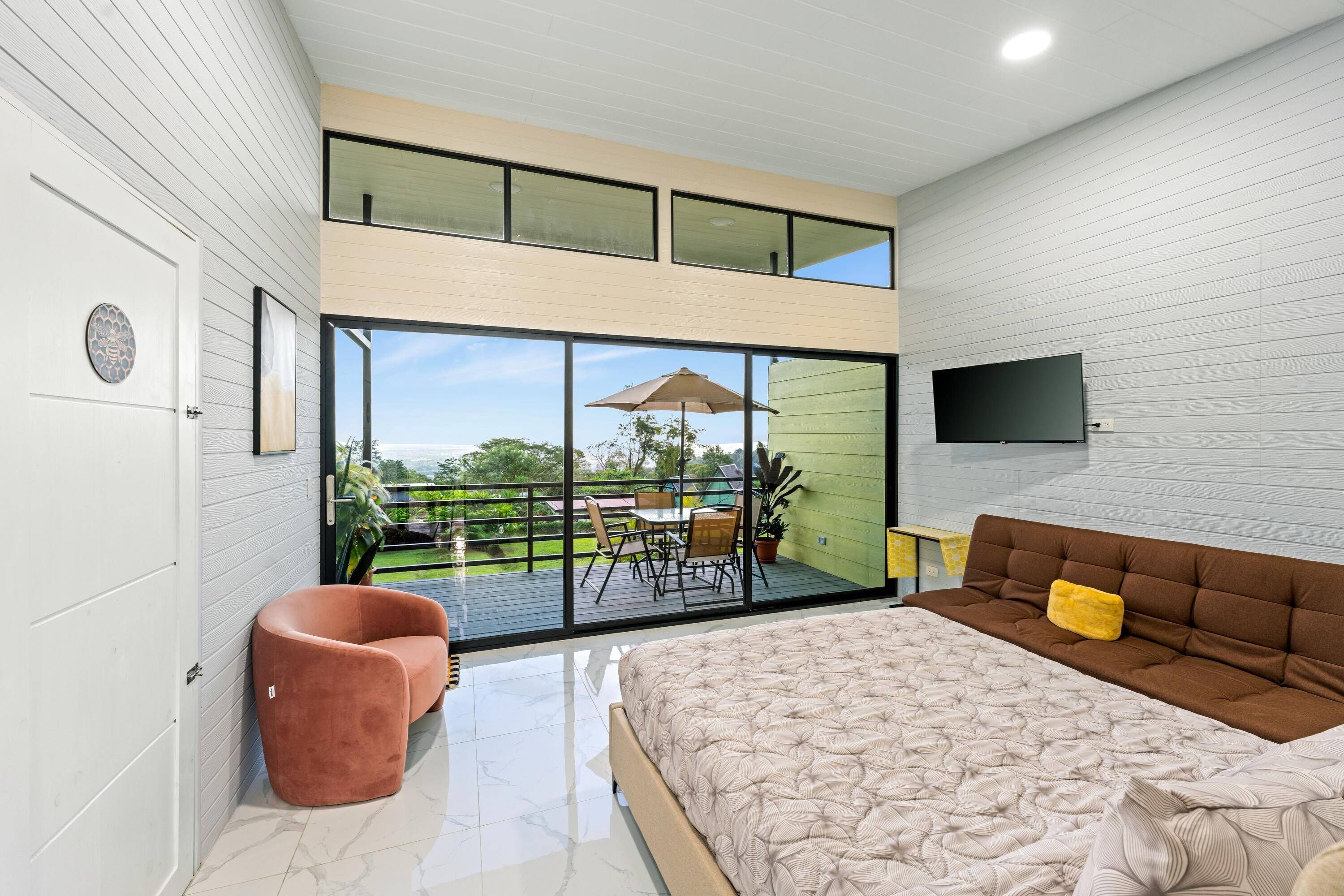 A modern bedroom with a large bed, a TV mounted on the wall, and a balcony with outdoor furniture.
