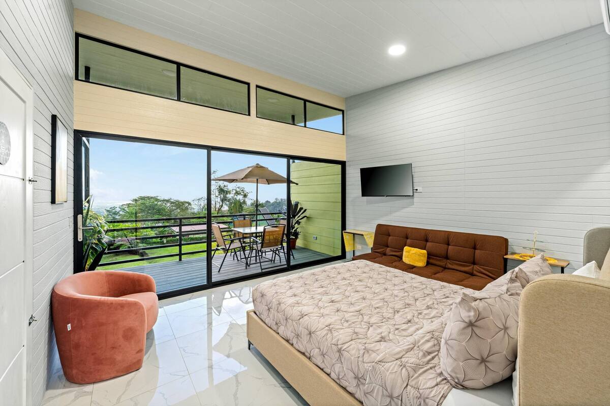 Un dormitorio moderno con una cama grande, un sofá, una silla y un balcón con mobiliario exterior.