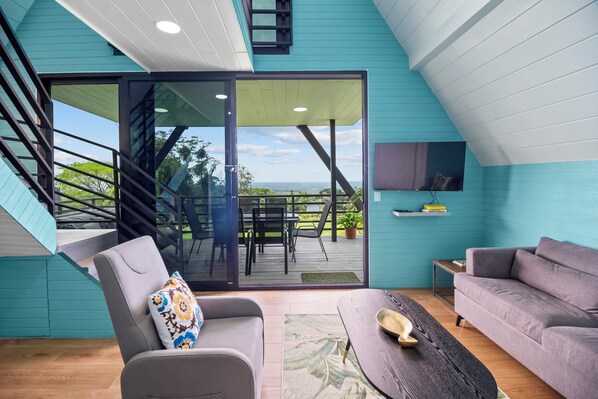 A modern interior with a grey sofa, a wooden coffee table, and a view through a glass door to an outdoor deck. - Junglo Resort and Spa (Peñas Blancas)