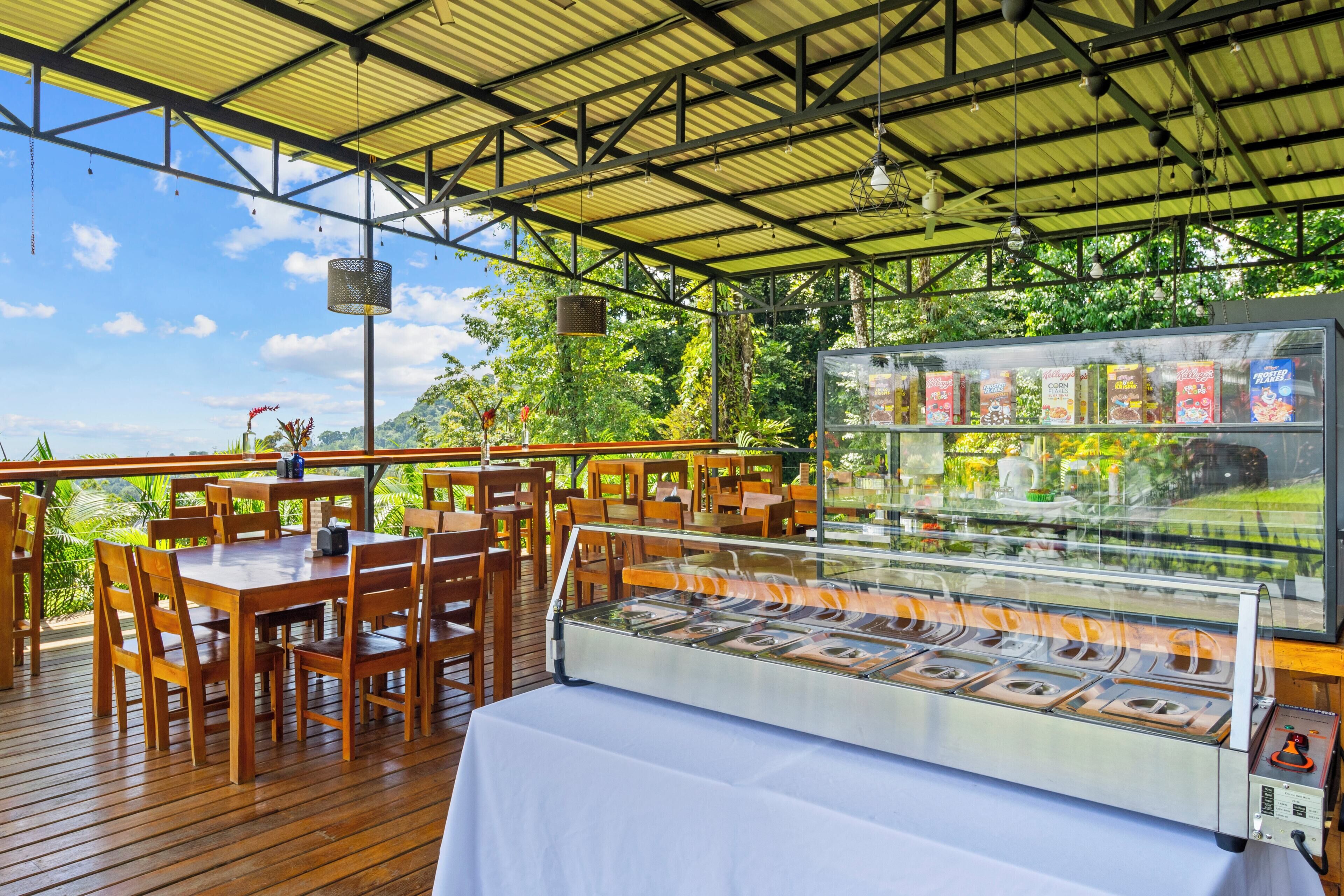 An outdoor dining area with wooden tables and chairs, a food display case, and a view of the surrounding greenery.