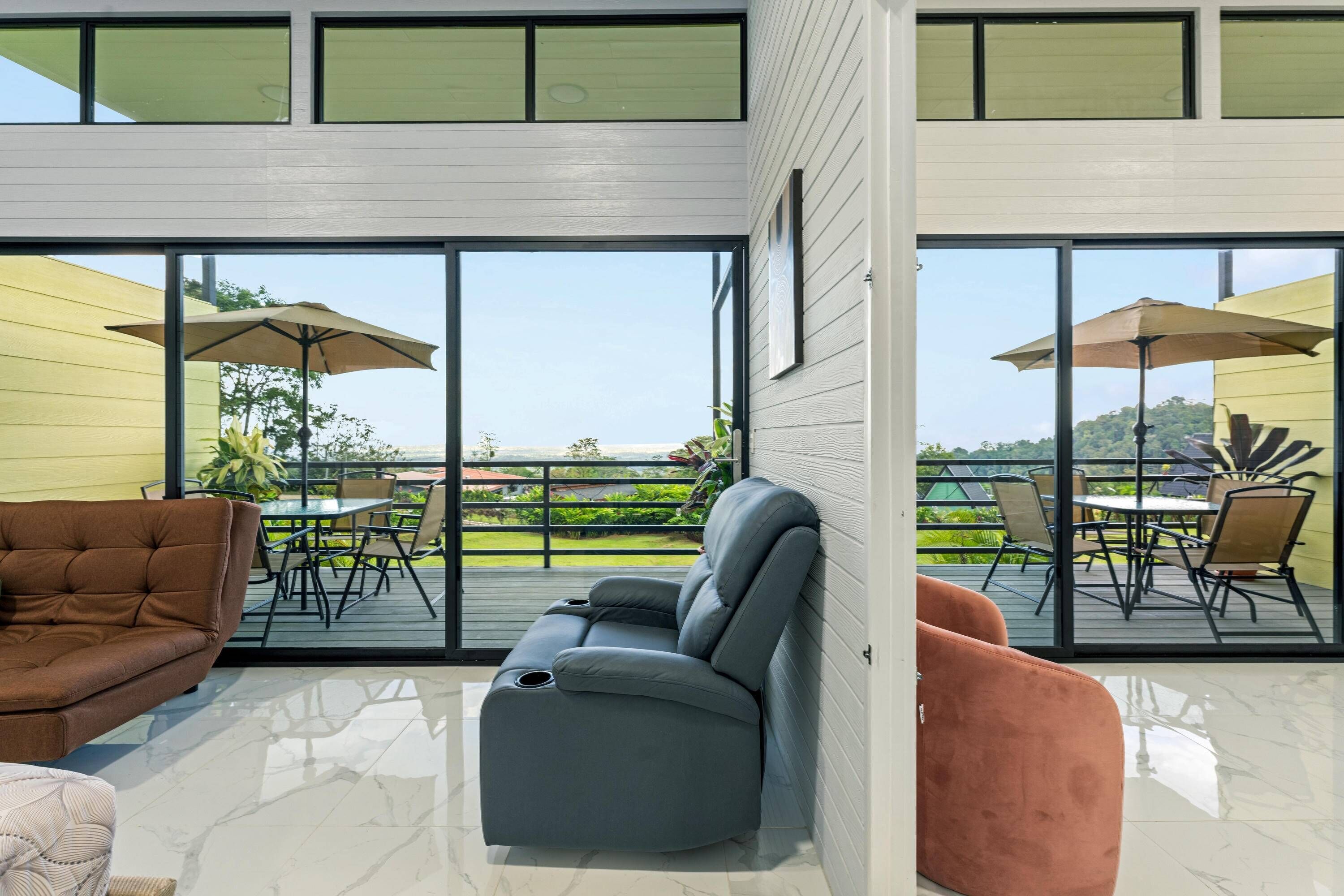 A modern living room with a grey recliner, a brown sofa, and a pink armchair. Large windows offer a view of a deck with outdoor furniture.