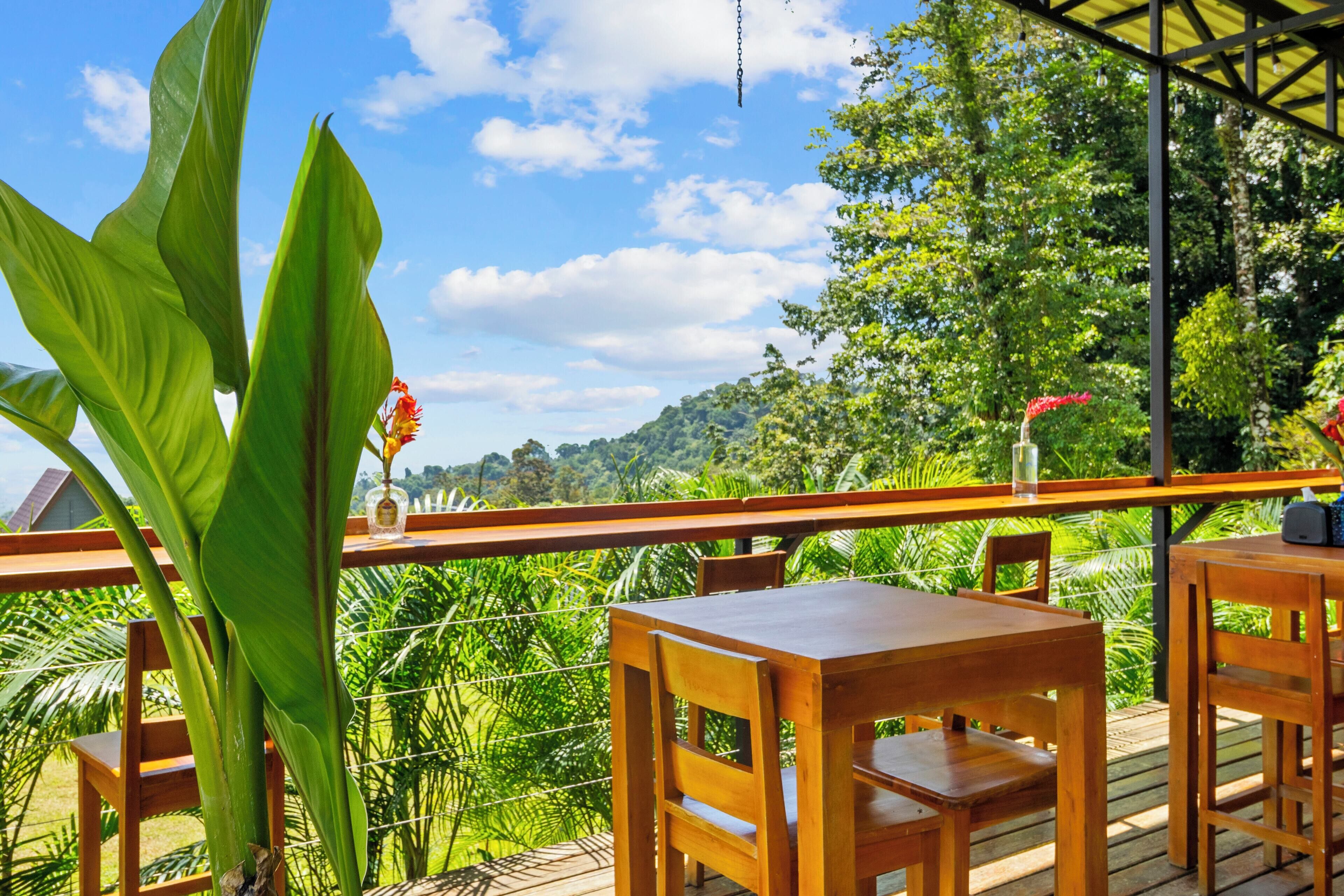 A wooden deck with tables and chairs, surrounded by lush greenery and a clear blue sky.
