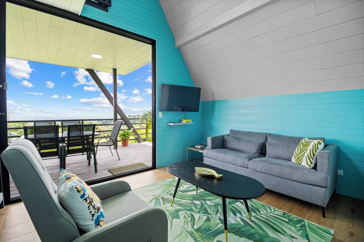 Una sala de estar moderna con un sofá gris, una mesa de centro negra y una alfombra verde estampada. Hay un televisor montado en la pared y una ventana que da a una terraza con mobiliario exterior.