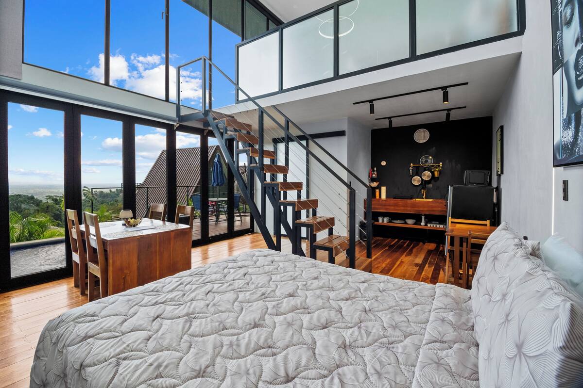Un dormitorio espacioso con una cama grande, una zona de comedor con una mesa y sillas, una escalera y vistas al exterior.