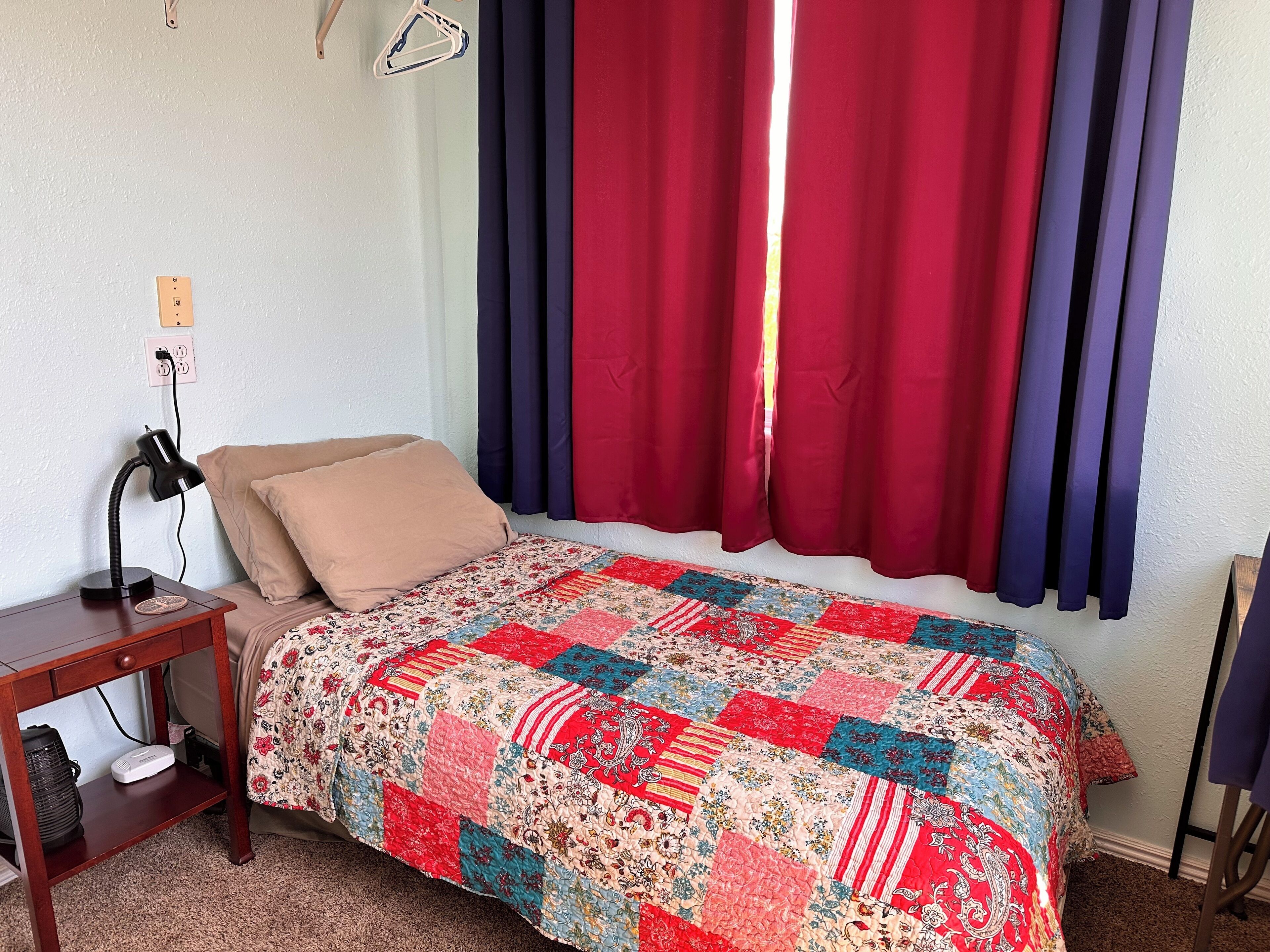 A single bed with a colorful quilt, a bedside table with a lamp, and a power outlet on the wall.