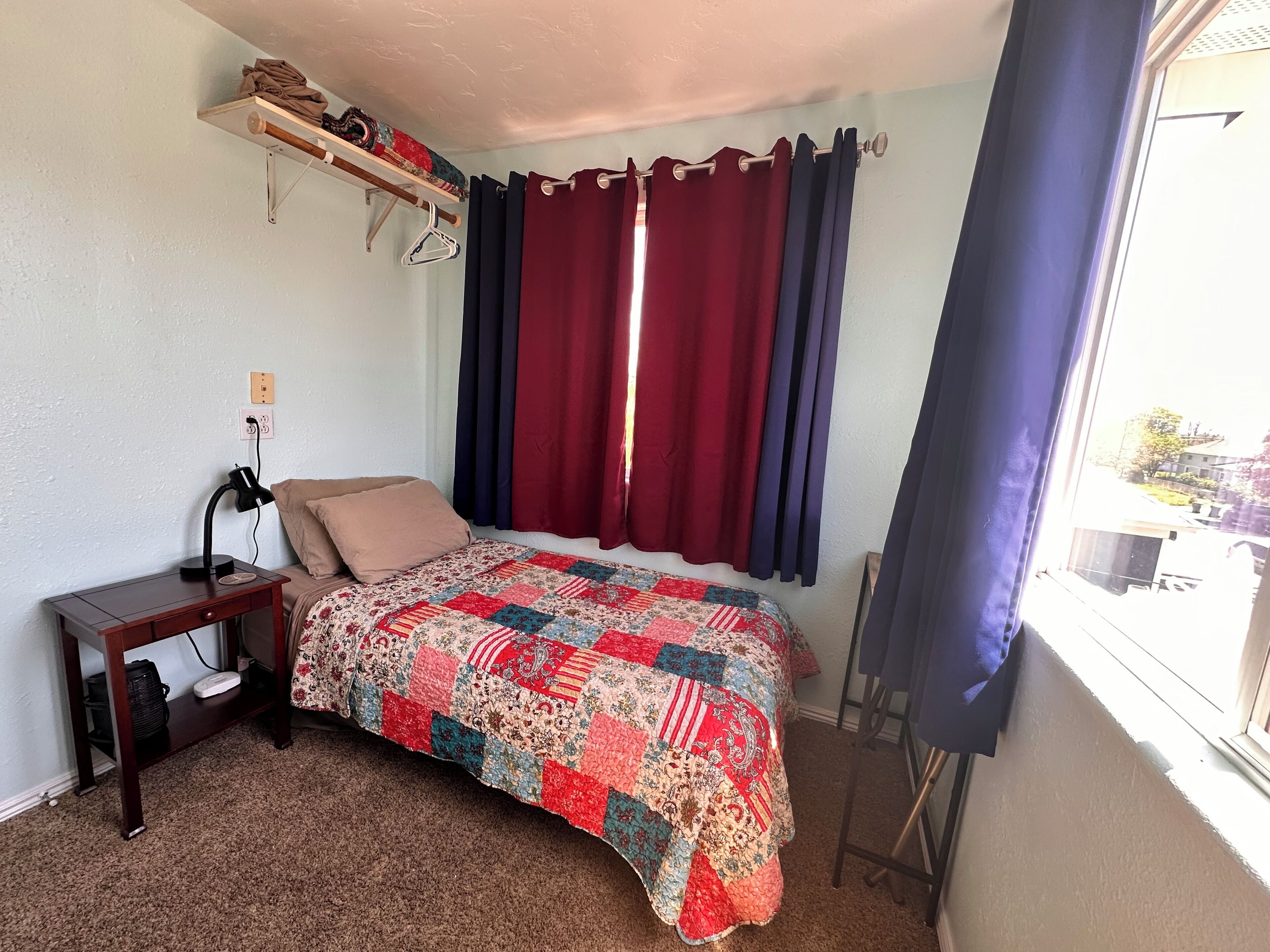 A bedroom with a bed, a nightstand, a lamp, a window with curtains, and a shelf with folded clothes.