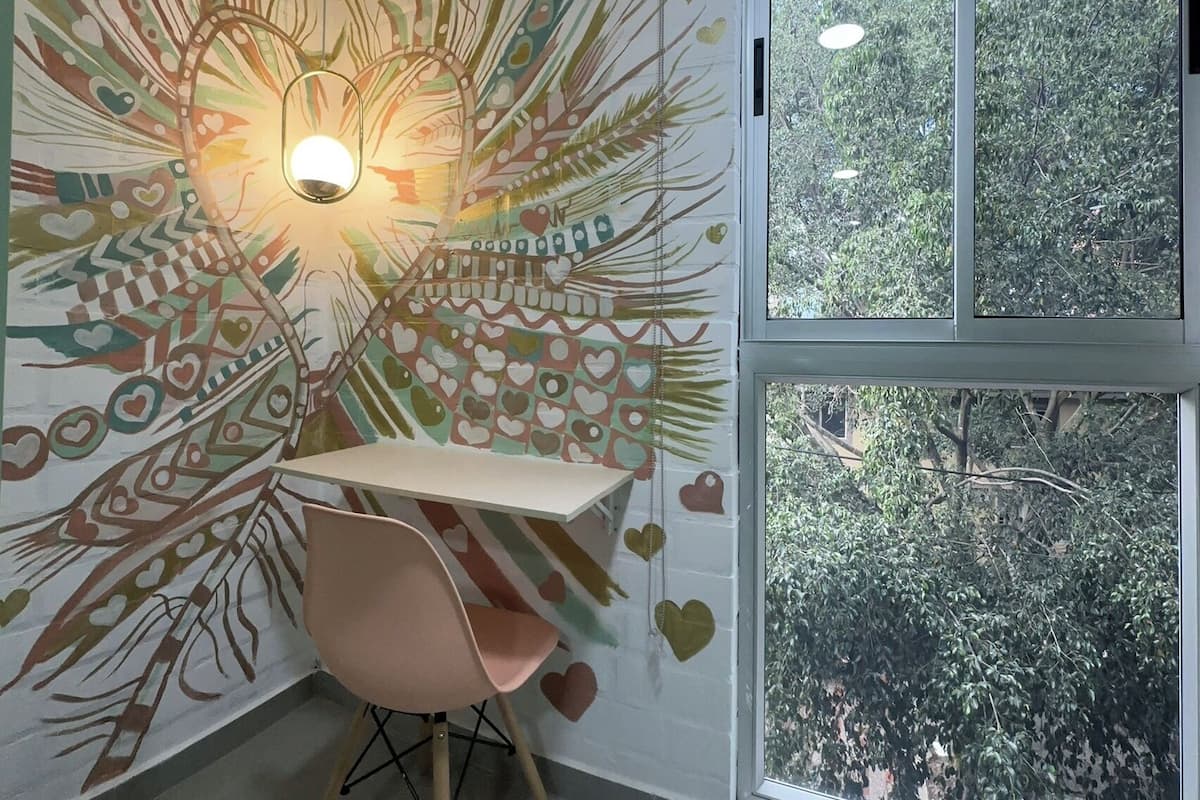 Une pièce d’angle avec une fresque murale, une table, une chaise et une grande fenêtre donnant sur les arbres.