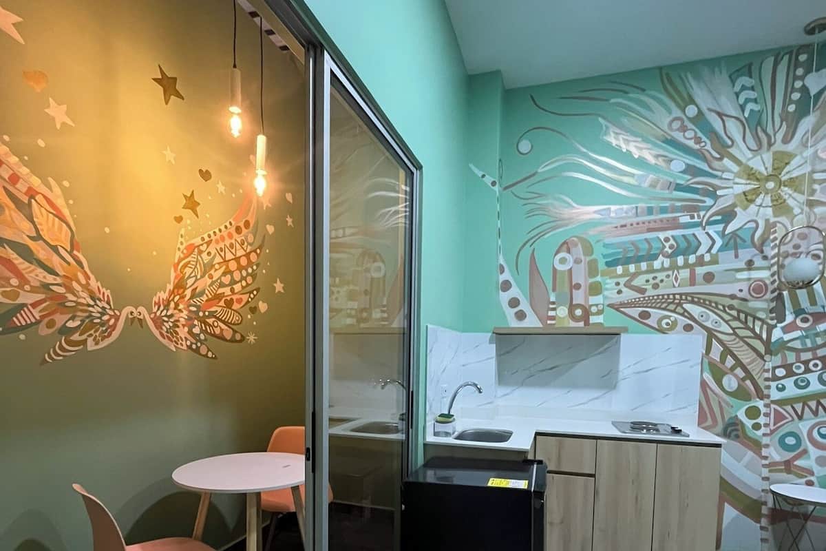 Une pièce avec une cloison en verre, une petite table avec des chaises, un réfrigérateur, un évier et un mur orné d’une fresque colorée.