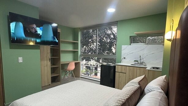 Ein Schlafzimmer mit Bett, einem an der Wand befestigten Fernseher, einem Fenster mit Blick auf Bäume und einer Küchenzeile.