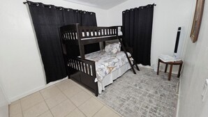 Une chambre avec un lit superposé, une petite table et une chaise, un radiateur et des rideaux noirs.