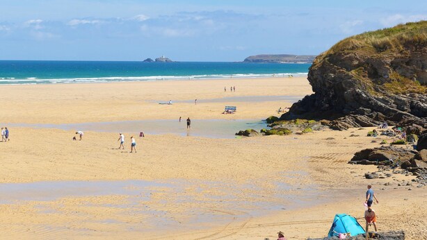 A wide sandy beach with a rocky outcrop on the right, a clear blue sky, and calm sea.
